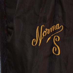 May include: Close-up of a dark brown garment with a satin-like texture. The garment features gold embroidered text that reads "Norma S". The lining has a dark fur trim.
