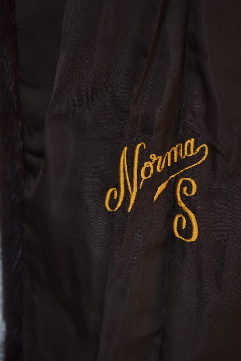 May include: Close-up of a dark brown garment with a satin-like texture. The garment features gold embroidered text that reads "Norma S". The lining has a dark fur trim.