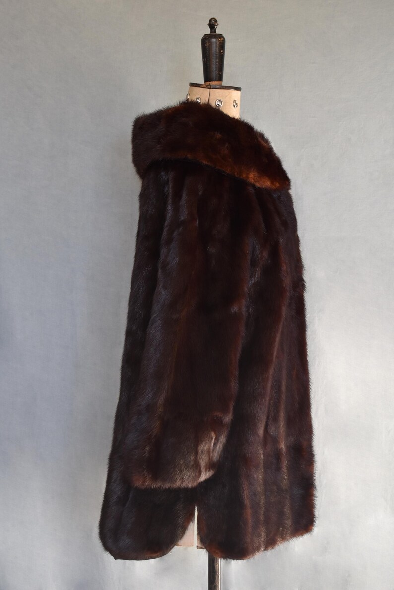 May include: A dark brown fur coat with a wide collar, displayed on a wooden mannequin. The coat has a long, flowing design and appears to be made of soft, luxurious fur. The coat is a rich, deep brown color.