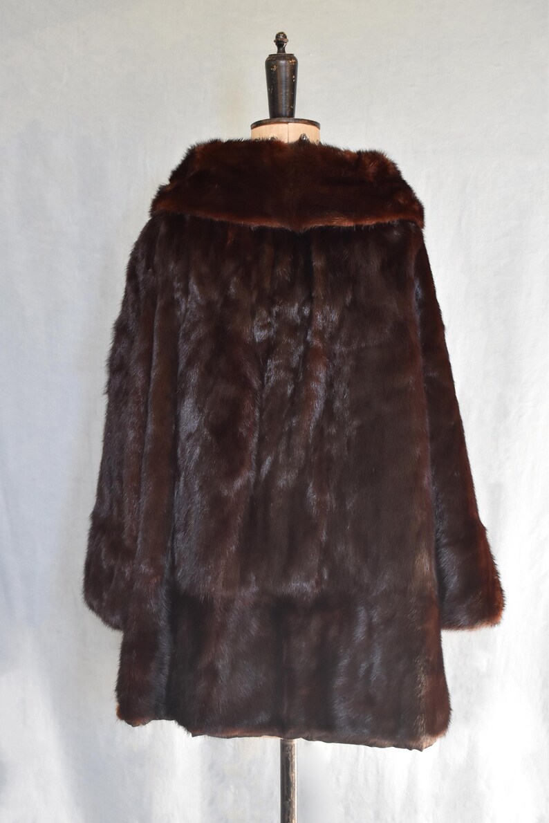 May include: A dark brown fur coat displayed on a mannequin. The coat has a wide collar and long sleeves. The fur appears dense and luxurious, with a rich, deep color. The coat is a classic outerwear garment.