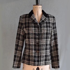 Puede incluir: Una chaqueta de cuadros en blanco y negro con cuello de terciopelo negro. La chaqueta tiene cierre de botones y mangas largas.