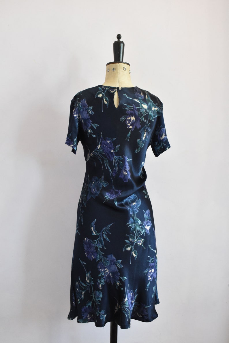 K&ouml;nnte beinhalten: Ein dunkelblaues Seidenkleid mit Blumenmuster. Das Kleid hat einen runden Ausschnitt und kurze &Auml;rmel. Das Kleid ist am Oberk&ouml;rper figurbetont und weitet sich zu einem leicht ausgestellten Rock.