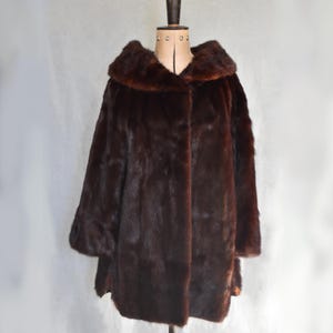 May include: A dark brown fur coat with a large collar and three-quarter sleeves. The coat is displayed on a vintage dress form against a plain white backdrop. The fur appears soft and luxurious.