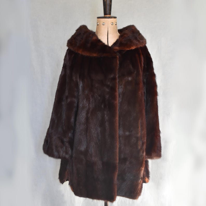 May include: A dark brown fur coat with a large collar and three-quarter sleeves. The coat is displayed on a vintage dress form against a plain white backdrop. The fur appears soft and luxurious.