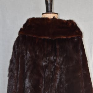 May include: A dark brown fur coat with a wide collar, displayed on a mannequin. The coat's fur appears soft and luxurious, with a rich, deep color. The mannequin is dark brown and has a wooden base.