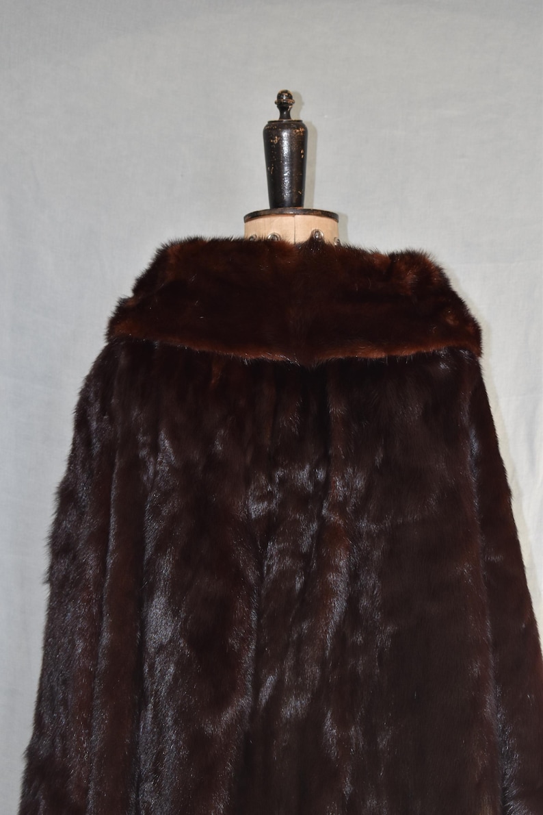 May include: A dark brown fur coat with a wide collar, displayed on a mannequin. The coat's fur appears soft and luxurious, with a rich, deep color. The mannequin is dark brown and has a wooden base.