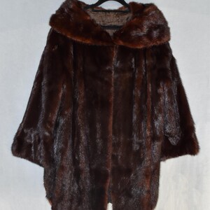 May include: A dark brown fur coat with a large collar and wide sleeves hangs on a black hanger. The coat has a smooth, luxurious texture and a classic design, perfect for cold weather. The coat is a rich, deep brown color.
