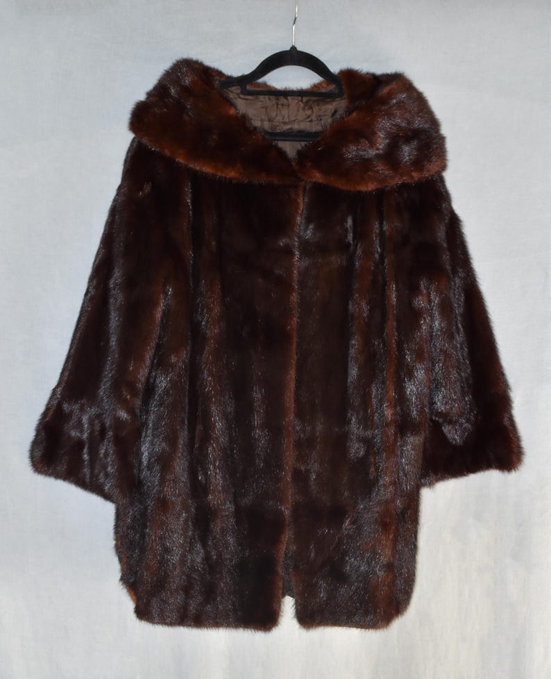 May include: A dark brown fur coat with a large collar and wide sleeves hangs on a black hanger. The coat has a smooth, luxurious texture and a classic design, perfect for cold weather. The coat is a rich, deep brown color.