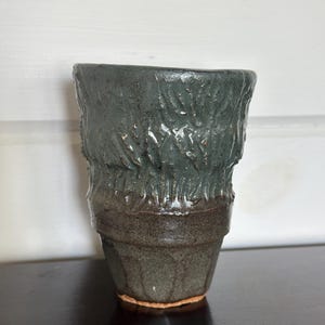 以下が含まれることがあります： テクスチャ加工された表面を持つ濃い緑色のセラミックマグカップ。マグカップは茶色の縁と底を持っています。