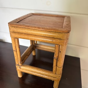 May include: A small, square bamboo side table with a slatted top. The table is constructed with bamboo poles and has a natural, light brown finish. The table is approximately 12 inches tall.
