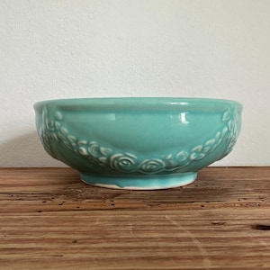 May include: A turquoise ceramic bowl with a floral design around the rim. The bowl is sitting on a wooden surface.