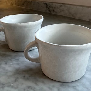 Pode incluir: Duas canecas de cerâmica branca com uma superfície texturizada. As canecas têm uma forma arredondada e uma única alça.