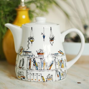 May include: White ceramic teapot featuring a black and white illustration of a bookshelf filled with books and hanging plants. The teapot has a spout and a handle.
