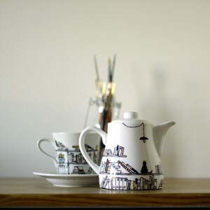 May include: White teapot and teacup with black and brown illustrations of bookshelves and a cat. The teapot has a black string with a lightbulb hanging from it.