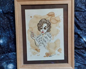 May include: Framed artwork featuring a caricature of a woman with red lipstick and a cigarette, set against coffee stain-like splatters. The woman wears a patterned blouse. The artwork is in a light brown wooden frame.