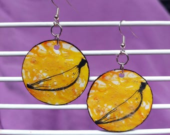 May include: A pair of circular dangle earrings featuring a yellow and white mottled background with a black line drawing of a banana on each. The earrings are attached to silver-toned metal fishhook wires and hang from small circular jump rings.