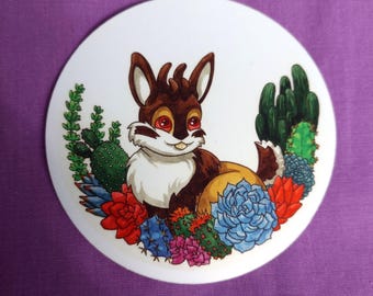 May include: A circular sticker featuring a brown and white cartoon creature with large red eyes, surrounded by a colorful arrangement of various green, blue, red, and pink succulents and cacti against a plain white background.