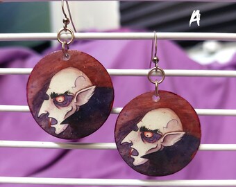 May include: A pair of circular drop earrings featuring a hand-painted illustration of a vampire with pale skin, dark eyebrows, and a single visible fang. The background of the circular discs is a deep red and purple gradient, and the letter A is visible.