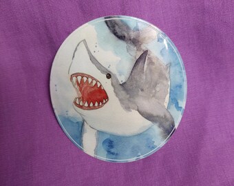 May include: A circular pin featuring a watercolor illustration of a shark with an open mouth showing sharp teeth. The shark is depicted in shades of gray and white against a blue, watery background with visible brushstroke textures.