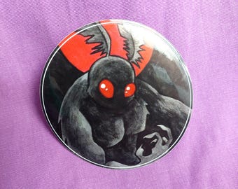 May include: A circular pinback button featuring a stylized illustration of a dark, furry creature with large, glowing red eyes and prominent red wing-like shapes behind its head, set against a deep black background with a thin white border.