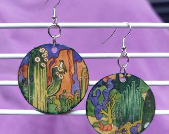 May include: A pair of circular dangle earrings featuring a desert landscape illustration. The left earring shows a tall cactus with a bird, while the right earring depicts various cacti and a small rabbit. Both have silver-toned metal hooks.