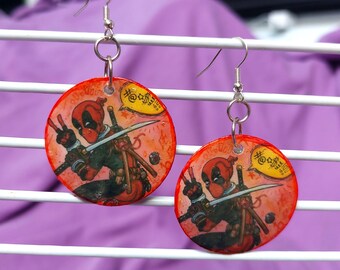 May include: A pair of round dangle earrings featuring a red and black comic book character illustration. The character holds a sword and makes a peace sign, with a yellow speech bubble containing symbols. The earrings have silver-toned metal hooks.