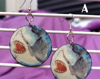 May include: A pair of circular dangle earrings featuring a watercolor-style illustration of a shark with an open mouth showing red teeth. The shark is depicted in shades of blue, gray, and white. The earrings have silver-toned metal hooks.