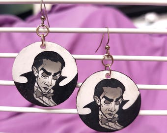 May include: A pair of circular dangle earrings featuring a black and white illustration of a vampire character with slicked-back hair, a high-collared cape, and a bowtie. The earrings hang from silver-toned metal hooks and circular jump rings.