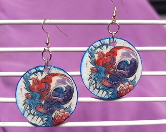 May include: A pair of circular dangle earrings featuring a hand-painted design of a human skull in shades of red, purple, and blue, accented with floral elements and sharp, thorn-like lines. The earrings hang from silver-toned metal hooks.