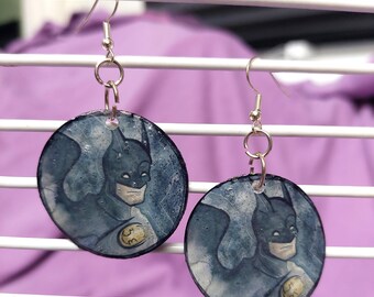 May include: A pair of circular dangle earrings featuring a graphic illustration of Batman in a dark blue and grey color palette. Each earring is suspended from a silver-toned metal hook and includes a small circular jump ring connector.