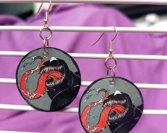 May include: A pair of circular dangle earrings featuring a graphic illustration of a black creature with a large, open mouth, sharp white teeth, and a long, winding red tongue against a grey background, suspended from silver-toned metal hooks.