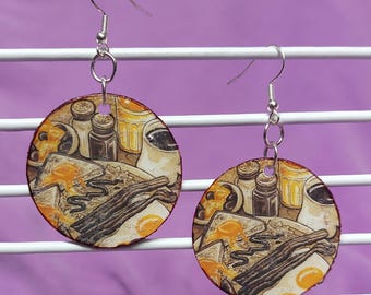 May include: A pair of round dangle earrings featuring a detailed illustration of a breakfast meal with bacon, eggs, toast, and coffee. The earrings are suspended from silver-toned metal hooks and display a warm color palette of brown, yellow, and white.