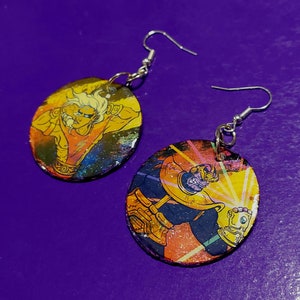 May include: A pair of earrings featuring round, colorful, painted images of comic book characters. The earrings are silver and have a dangling design.