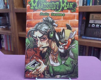May include: A comic book titled The Makeshift Man 07 by JoJo Seames. The cover features a stylized illustration of a person with long dark hair, wearing green headphones and a green jacket, holding a green cassette player against a brick wall.