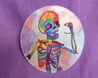 May include: A circular pin featuring a colorful watercolor illustration of a skeleton. The skeleton is depicted with a vibrant rainbow palette, including shades of red, orange, yellow, green, blue, and purple, set against a soft, mottled background.