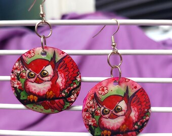 May include: A pair of circular dangle earrings featuring a vibrant red owl illustration perched on a branch amidst pink cherry blossoms. The earrings are suspended from silver-toned metal hooks and connected by small circular jump rings.