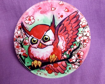 May include: A circular sticker featuring a red owl with large yellow eyes perched on a dark branch. The owl is surrounded by pink cherry blossoms and green foliage against a watercolor background. A small cluster of red cherries hangs from the branch.