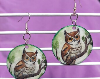 May include: A pair of round dangle earrings featuring a hand-painted illustration of a brown and white owl perched on a tree branch. The circular discs have a green border and are attached to silver-toned metal fishhook ear wires.
