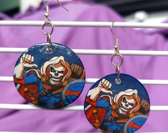 May include: A pair of circular dangle earrings featuring a graphic illustration of a skeleton character wearing a blue and orange outfit with a cape, hood, and shield. The earrings are suspended from silver-toned metal fishhook ear wires.