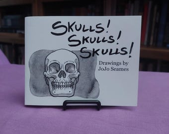 May include: A white rectangular booklet titled Skulls! Skulls! Skulls! with a black ink drawing of a human skull centered on the cover. The text Drawings by JoJo Seames is printed in black on the right side. The booklet rests on a black stand.