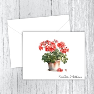 May include: A white card with a watercolor illustration of a potted geranium plant with pink flowers. The card has the name "Kathleen Mathews" written at the bottom.