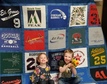 May include: A blue and white quilt made from t-shirts with various sports team logos and names, including "Elk Grove Babe Ruth", "Cougar Baseball", "All Stars", "Athletics", and "Sacramento Heat". Two children are sitting on the quilt, one holding a toy gun.