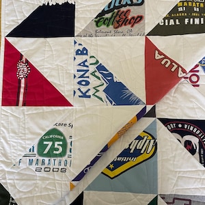 May include: A handmade quilt composed of various t-shirt designs and white fabric squares. The quilt features a patchwork design with triangular and square shapes. The t-shirt designs include text and graphics, with colors like red, blue, and green.