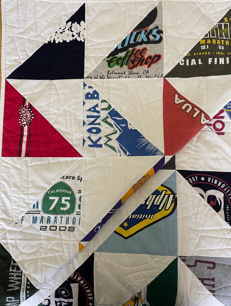 May include: A handmade quilt composed of various t-shirt designs and white fabric squares. The quilt features a patchwork design with triangular and square shapes. The t-shirt designs include text and graphics, with colors like red, blue, and green.