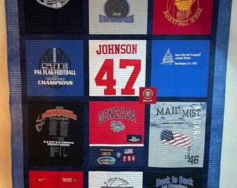 May include: A blue quilt with various patches of different colored shirts. The shirts have logos and text, including "SJE", "Johnson 47", "Gonzaga", "Maid Mist", "Little Dribblers", and "Back to Back Pal Champions".