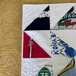 May include: A handmade quilt with a white background and triangular patches of various colors and patterns. Some patches feature text and graphics, including "California 75" and "KONAB". The quilt is quilted with white thread.