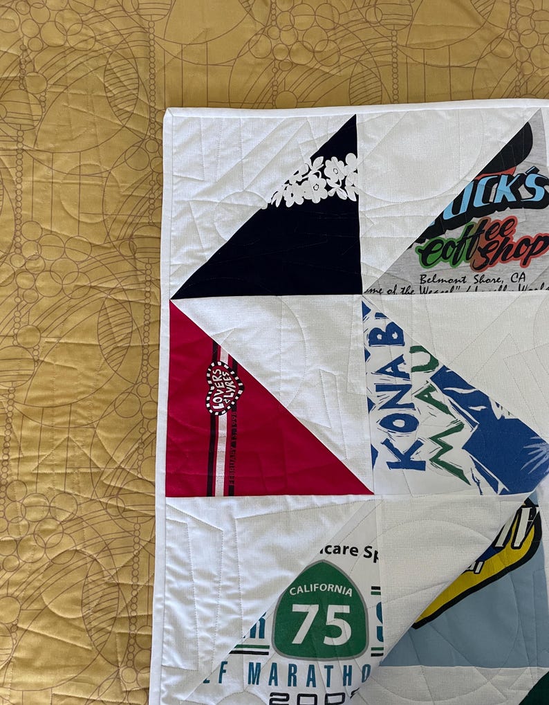 May include: A handmade quilt with a white background and triangular patches of various colors and patterns. Some patches feature text and graphics, including "California 75" and "KONAB". The quilt is quilted with white thread.