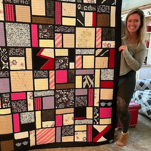 May include: A patchwork quilt with a black border and a variety of patterned squares in shades of pink, cream, black, and purple. The quilt features floral, geometric, and abstract designs, creating a visually dynamic textile.