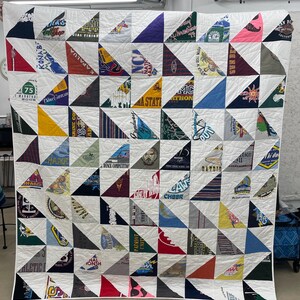 May include: A handmade quilt composed of triangular patches of various colors and patterns, including text and logos from t-shirts. The quilt is sewn together with white fabric and features a grid-like pattern. The overall design is a patchwork of memories.