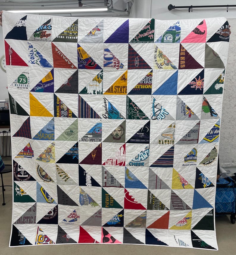 May include: A handmade quilt composed of triangular patches of various colors and patterns, including text and logos from t-shirts. The quilt is sewn together with white fabric and features a grid-like pattern. The overall design is a patchwork of memories.